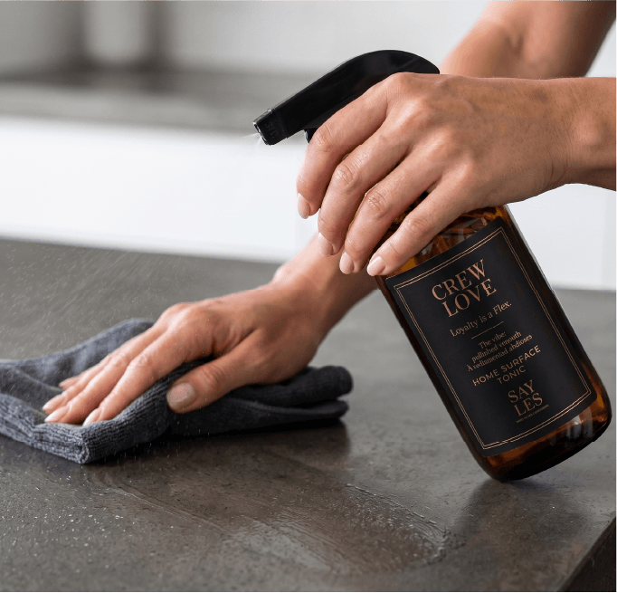 Close-up of hands wiping a dark countertop with a grey cloth while holding a bottle of Crew Love Home Surface Tonic.
