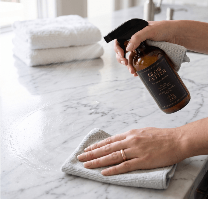 Close-up of a person spraying and wiping a white marble countertop with Glow Getter Home Surface Tonic using a microfiber cloth.